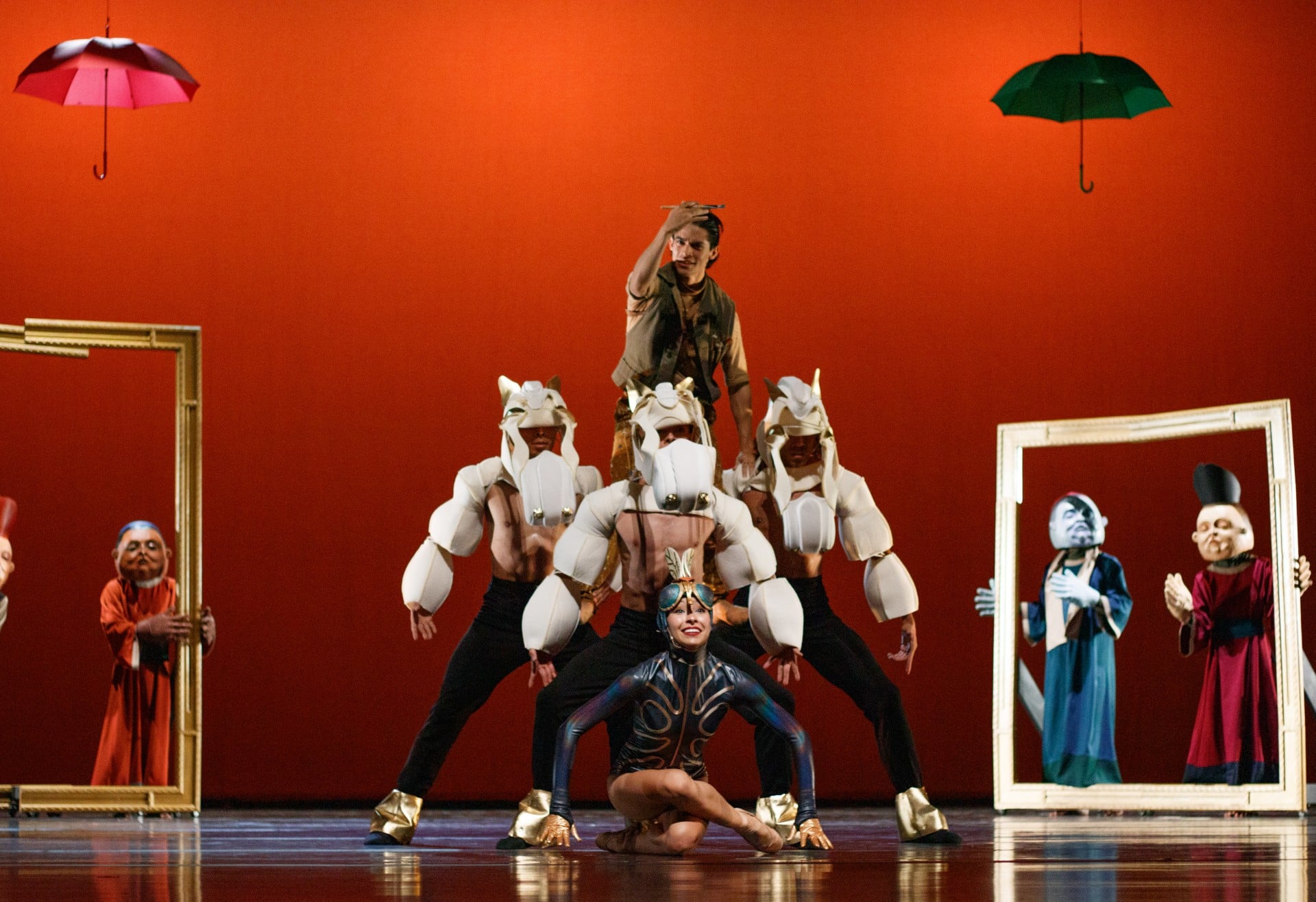 Una escena teatral vibrante con un fondo naranja intenso muestra a artistas en trajes surrealistas. Un performer en trajes azules está al frente, mientras otros con cabezas de caballos blancos y guantes dorados lo rodean. Marcos dorados y personajes con máscaras completan la composición.
